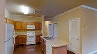 This single-floor residence is designed with functionality and comfort in mind. The kitchen serves as a central highlight, showcasing warm honey-toned wood cabinetry that lines the walls and provides ample storage space. White appliances, including a range with oven, built-in microwave, and dishwasher, complement the neutral countertops throughout. A peninsula extends into the kitchen space, housing a double-basin sink and offering additional prep surface area, making it ideal for everyday cooking and entertaining. Crown molding runs along the ceiling, adding a refined architectural detail that elevates the overall aesthetic. The flooring features dark hardwood, lending warmth and a classic feel to the space. Overhead lighting keeps the kitchen well-illuminated, while a bi-fold door closet nearby adds convenient storage access. The layout allows for easy movement between the kitchen and adjoining areas, with doorways visible in the background that connect to additional rooms. The warm yellow-toned walls tie the space together, creating a cozy and welcoming atmosphere throughout this well-appointed single-level home.