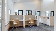 This single-floor amenity space is thoughtfully designed to support both focused work and casual interaction. The interior features soaring ceilings that lend an open, airy quality throughout, with crisp white walls and patterned carpet tile flooring creating a clean, contemporary aesthetic.

The co-working area offers multiple built-in desk stations separated by low dividing panels, each equipped with power outlets and topped with light-toned surfaces. Rattan-backed task chairs complement the coastal-inspired decor, while decorative woven vessels displayed above the partitions add warmth. Black-framed transom windows above the stations allow natural light to filter in while maintaining privacy.

A glass-enclosed private office, framed in bold black steel, provides a quiet workspace for focused tasks, outfitted with a desk, task chair, and pendant lighting. Large shuttered windows offer views of the surrounding outdoor amenities.

A lounge area features rich leather accent chairs paired with a natural wood side table and a wall-mounted display screen — ideal for informal meetings or relaxation. Adjacent to the lounge, full-height glass and white-framed double doors open directly to the outdoors, flooding the entry with natural light and providing seamless access to exterior grounds.

A built-in service counter with white shaker cabinetry, stone countertops, and matte black hardware completes the space, offering a functional administrative or reception point.