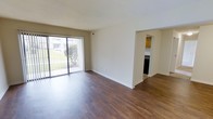 This single-floor space offers a clean, open layout highlighted by rich, dark hardwood-style flooring that extends throughout the main living area, creating a warm and grounded aesthetic. The living room is generously proportioned, with neutral beige walls and crisp white trim that lend a bright, airy feel. A large sliding glass door fitted with vertical blinds anchors one wall, allowing abundant natural light to pour in while providing direct views of a landscaped outdoor area. The seamless connection to the outdoors enhances the overall sense of space and openness.

A defined opening along the far wall leads into the kitchen, which is partially visible from the living area. The kitchen features wood-toned cabinetry and appears to include dark appliances, offering a functional and organized cooking space. The transition between rooms is clearly delineated, maintaining a structured flow throughout the unit.

Overall, the space presents a straightforward and practical layout with an emphasis on natural light, durable flooring, and a neutral color palette that provides a versatile backdrop for a variety of interior styles.
