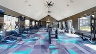 The Prism At Dimond Ridge Fitness Center is a state-of-the-art facility designed to cater to all fitness needs. Spanning 1600 square feet on a single floor, this gym boasts a striking architectural design with vaulted ceilings that create an open, airy atmosphere. The space is illuminated by a combination of natural light streaming through large windows and strategically placed recessed lighting, complemented by unique ceiling fans that add both functionality and style.

The gym's most eye-catching feature is its vibrant geometric flooring, combining shades of blue, teal, and gray in an abstract pattern that energizes the space. This modern aesthetic is balanced by sleek charcoal walls, creating a contemporary and motivating environment for exercise.

Equipment-wise, the center is well-equipped with a diverse range of modern fitness machines. Cardio enthusiasts will find multiple treadmills, ellipticals, and stationary bikes, while strength training options include weight machines, free weights, and dedicated areas for resistance exercises. The layout is thoughtfully designed to allow for easy navigation and efficient use of space.

Additional amenities include wall-mounted TVs for entertainment during workouts, mirrored walls for form checking, and storage solutions for personal belongings. The facility also features glass doors leading to an outdoor area, providing options for open-air exercises or cool-down sessions.

Overall, the Prism At Dimond Ridge Fitness Center offers a comprehensive and visually appealing workout space that caters to a variety of fitness preferences and levels.