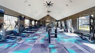 The Prism At Dimond Ridge Fitness Center is a state-of-the-art facility designed to cater to all fitness needs. Spanning 1600 square feet on a single floor, this gym boasts a striking architectural design with vaulted ceilings that create an open, airy atmosphere. The space is illuminated by a combination of natural light streaming through large windows and strategically placed recessed lighting, complemented by unique ceiling fans that add both functionality and style.

The gym's most eye-catching feature is its vibrant geometric flooring, combining shades of blue, teal, and gray in an abstract pattern that energizes the space. This modern aesthetic is balanced by sleek charcoal walls, creating a contemporary and motivating environment for exercise.

Equipment-wise, the center is well-equipped with a diverse range of modern fitness machines. Cardio enthusiasts will find multiple treadmills, ellipticals, and stationary bikes, while strength training options include weight machines, free weights, and dedicated areas for resistance exercises. The layout is thoughtfully designed to allow for easy navigation and efficient use of space.

Additional amenities include wall-mounted TVs for entertainment during workouts, mirrored walls for form checking, and storage solutions for personal belongings. The facility also features glass doors leading to an outdoor area, providing options for open-air exercises or cool-down sessions.

Overall, the Prism At Dimond Ridge Fitness Center offers a comprehensive and visually appealing workout space that caters to a variety of fitness preferences and levels.