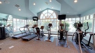 This single-floor fitness center is thoughtfully designed to provide a bright, welcoming environment for exercise. The space is defined by its striking vaulted ceilings and a large arched feature window, which floods the interior with natural light and frames views of the surrounding greenery. Soft blue-painted walls and neutral carpeted flooring contribute to a calm, focused atmosphere throughout.

The cardio zone is well-appointed with multiple treadmills, upright and recumbent stationary bikes, and elliptical cross-trainers, each positioned to take advantage of the expansive window views. Wall-mounted flat-screen televisions are strategically placed to keep users engaged during workouts. A dedicated strength training area occupies a separate section of the room, outfitted with a multi-station cable machine, adjustable weight benches, and a full dumbbell rack. Floor-to-ceiling mirrors line the strength area walls, enhancing the sense of space and allowing users to monitor their form.

Recessed lighting supplements the generous natural light, ensuring the space is well-illuminated at all times. A water station and storage shelving are conveniently accessible near the entrance, and a glazed exterior door provides direct access to the outdoors. The overall layout is open and uncluttered, offering ample room to move freely between equipment stations.