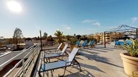 This rooftop terrace offers a well-appointed open-air space designed for relaxation and leisure with sweeping panoramic views of the surrounding urban landscape, including a distant city skyline framed by palm trees and clear blue skies. The terrace is finished with large-format stone-toned tiles that lend a clean, durable surface throughout the deck area. Multiple adjustable lounge chairs in white and blue are arranged across the space, providing ample seating for sunbathing or simply taking in the views. Decorative black lamp posts are positioned across the terrace, ensuring the space is well-lit during evening hours. Potted tropical plants, including a striking banana tree, add lush greenery and a resort-like ambiance to the setting. A low-profile white metal railing runs along the perimeter, maintaining an unobstructed sightline across the rooftop. Access to the terrace is provided via a stone-tiled staircase with a handrail, flanked by white stucco walls that reflect the warm Southern California architectural aesthetic. The overall design of the terrace balances functionality with a relaxed, sun-soaked atmosphere, making it a standout amenity with commanding views in every direction.
