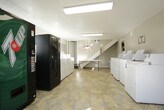 This shared laundry facility is designed to serve a community with efficiency and convenience in mind. The space is spread across a single floor and is organized into interconnected areas, each serving a distinct function within the overall laundry experience. The flooring throughout features a durable stone-look vinyl tile that is both practical and easy to maintain, complementing the neutral gray and white wall tones that lend the space a clean, functional aesthetic. Overhead fluorescent lighting ensures the entire facility is well-lit throughout.

The main laundry area is generously equipped with multiple top-loading washing machines and a bank of stacked dryers, providing ample capacity for high-demand use. Folding tables are conveniently positioned nearby, offering residents a dedicated space to sort and fold laundry. A connected secondary room houses additional washers and dryers, expanding the facility's overall capacity.

A casual seating area features a small table and chairs, giving residents a comfortable place to wait. Vending machines are also available on-site for added convenience. Large windows in the seating area allow natural light to filter in, creating a bright and welcoming atmosphere. The textured ceiling and well-maintained walls contribute to the facility's tidy, community-oriented environment.