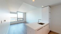 This single-floor unit showcases a clean, contemporary design with a cohesive all-white palette and warm wood-tone flooring that runs throughout the space. The open-concept living and kitchen area forms the heart of the unit, where large floor-to-ceiling windows frame panoramic views of the surrounding cityscape and waterway, flooding the space with natural light.

The kitchen is outfitted with flat-front white cabinetry, a white quartz island with an undermount sink, and stainless steel appliances including a gas cooktop, wall oven, and refrigerator. Recessed lighting adds a polished, functional finish to the space.

The living area is generously proportioned, with wide-format windows continuing the view-centric design. Each bedroom features its own large window with elevated city views and hardwood-style flooring. The bathroom presents a refined aesthetic with a dual-basin white quartz vanity, tall white cabinetry, chrome fixtures, and tile flooring.

A dedicated laundry area houses a stacked full-size washer and dryer. A clean entry corridor with a bold dark front door provides a defined arrival sequence. Throughout, the design prioritizes natural light, open sightlines, and a minimalist modern sensibility.