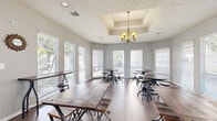 Located at 15215 Blue Ash Dr in Houston, TX, this inviting single-floor gathering space offers a warm and functional environment designed for community interaction and social engagement. The layout comprises two open rooms that flow cohesively, unified by rich hardwood flooring and soft gray walls throughout.

The main gathering area makes a strong architectural impression with soaring ceilings, exposed brick columns, and clerestory windows that draw in abundant natural light. A wall-mounted television and multiple seating arrangements — including rustic wood communal tables with industrial-style benches and adjustable stools — make this area well-suited for group gatherings or casual meetings.

An adjoining sunlit room features a distinctive tray ceiling adorned with an elegant brass chandelier, creating a refined yet relaxed atmosphere. Floor-to-ceiling windows on multiple walls flood the space with natural light and offer views of surrounding greenery. Smaller round tables with adjustable stools are arranged throughout, providing intimate seating options.

Together, the two rooms balance rustic charm with contemporary functionality, highlighted by decorative wall accents and thoughtful lighting. Glass-paneled doors connect the interior spaces, enhancing openness and visual flow. This well-appointed space offers a welcoming atmosphere ideal for residents and guests alike.