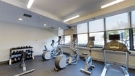 Located at 3 Sundial in Manchester, New Hampshire, this fitness center offers a well-appointed, single-floor exercise space designed to support an active lifestyle. The room is outfitted with a variety of modern cardio machines, including ellipticals, stationary bikes, and stair steppers, each equipped with individual display monitors for entertainment or workout tracking. The flooring features durable, dark rubberized material suited for high-traffic athletic use. Overhead, a combination of recessed lighting and linear fluorescent fixtures ensures the space is evenly and brightly illuminated throughout. A standout feature of the fitness center is its expansive floor-to-ceiling windows along one wall, which flood the room with natural light and provide open views of the surrounding outdoor environment. Roller shades offer the option to control light levels as needed. The walls are finished in a clean, neutral white, contributing to an airy and modern aesthetic. The ceiling incorporates visible HVAC vents, reflecting a practical, climate-controlled environment. Overall, the space presents a clean, functional, and modern fitness facility well-suited for daily exercise routines.