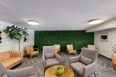 Located at 8750 Georgia Ave in Silver Spring, Maryland, this thoughtfully designed lounge offers a comfortable and inviting communal gathering space. The single-floor layout is anchored by a standout living area that blends modern aesthetics with mid-century inspired furnishings.

The lounge features a mix of seating options, including a tufted caramel leather sofa, warm wood-framed leather accent chairs, and plush gray upholstered armchairs arranged around a gold-toned circular coffee table. This variety of seating creates multiple conversation areas ideal for both relaxed socializing and casual meetings.

A dramatic floor-to-ceiling living green wall serves as the room's defining architectural feature, introducing a bold natural element that contrasts beautifully against the neutral walls and textured carpet flooring. Tall fiddle-leaf fig trees and potted plants are placed throughout, reinforcing the biophilic design theme.

Large floor-to-ceiling glass windows and glazed partition walls allow generous natural light to pour into the space, offering views of the surrounding greenery and maintaining a bright, open atmosphere. Circular flush-mount ceiling lights provide warm, even ambient illumination. Abstract wall art and carefully selected decorative accents complete the polished, contemporary look of this well-appointed lounge.