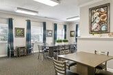 This single-floor event center offers a thoughtfully designed layout suited for gatherings, meetings, and private functions. The space is organized into several distinct areas that work together to support a complete event experience. Upon entry, a pass-through counter separates the main gathering area from the kitchen, providing a practical service interface. The fully equipped kitchen features stainless steel appliances, including a full-size refrigerator and microwave, white cabinetry with dark granite-style countertops, and tile flooring — offering ample preparation space for catering needs. An adjacent catering or serving room showcases a striking floor-to-ceiling mosaic tile accent wall, extended granite-topped white cabinetry, and tile flooring, providing generous buffet or setup space. The main gathering area is carpeted and accommodates multiple dining tables and chairs arranged for group seating. Large windows dressed with teal grommet-panel drapes allow natural light to fill the room, complemented by framed artwork on neutral-toned walls and crown molding details. Flush-mounted ceiling fixtures provide consistent overhead lighting throughout. The overall ambiance is clean, contemporary, and welcoming, making this event center a versatile and functional venue.