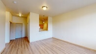 Located at 1341 NE Park Ln in Fairview, Oregon, this one-bedroom, one-bathroom unit offers a well-organized single-floor layout designed for comfortable everyday living. The entrance opens into a combined living and dining area featuring light-toned hardwood-style flooring, neutral cream walls, and a wall-mounted HVAC unit beneath a bright window that brings in natural light and outdoor views of the surrounding greenery. A flush-mount ceiling fixture and clean baseboard trim complete the polished look of this open area. The kitchen is designed as a galley-style space with warm honey-toned wood cabinetry lining both sides, light countertops with a tiled backsplash, and white appliances including a refrigerator, electric range, microwave, and dishwasher. A pass-through window connects the kitchen to the living area, enhancing the open feel. Conveniently tucked off the kitchen is an in-unit laundry area with a stacked washer and dryer. The bedroom is carpeted for comfort, features a ceiling fan, a window with outdoor views, and a spacious closet with a full-width hanging rod. The bathroom includes a combined tub and shower, light wood-style flooring, wood vanity cabinetry, and accessibility-friendly grab bars.