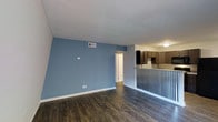 This single-floor apartment at Clifton Ridge Apartments offers an open-concept layout that seamlessly connects the living area to the kitchen. The living space is defined by striking blue accent walls contrasted against crisp white trim and ceiling, creating a clean, modern aesthetic. Dark hardwood-style flooring runs throughout, adding warmth and visual continuity across the space.

A half-wall partition with vertical metal spindles separates the living area from the kitchen, maintaining an open feel while providing subtle definition between the two spaces. The kitchen features dark espresso-toned cabinetry paired with a decorative tile backsplash, offering both style and functionality. Light countertops provide contrast against the darker elements, and a ceiling-mounted light fixture ensures adequate illumination.

A hallway visible from the main living area leads to additional rooms within the unit. Overhead ventilation is provided via ceiling-mounted HVAC vents, ensuring climate comfort throughout. The overall ambiance is contemporary and well-appointed, combining practical design elements with thoughtful aesthetic details for a cohesive living environment.