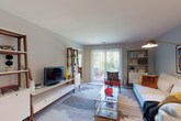 Located at 100 Conestoga Dr. in Marlton, NJ, this single-floor residence offers a well-organized layout with distinct living, dining, kitchen, sleeping, and bathroom areas. The living room is bright and open, featuring large sliding glass doors with vertical blinds that provide access to an outdoor patio and frame lush greenery views. The space accommodates a full seating arrangement centered around a glass coffee table, complemented by open wood-and-white shelving units on either side. The kitchen is efficiently designed with light maple cabinetry, dark granite-style countertops, hardwood-style flooring, and a full suite of white appliances including a refrigerator, range, microwave, and dishwasher. A ceiling fan overhead adds to the functionality. Adjacent to the kitchen, a dedicated dining area offers comfortable space for everyday meals. The bedroom is generously sized with plush carpeting, a bold navy accent wall, and windows that invite natural light. A walk-in closet provides substantial storage, and a full bathroom is conveniently accessible from the bedroom. The bathroom features tile flooring, a white vanity with ample counter space, and a shower-tub combination. Overall, the residence presents a practical and comfortable living environment throughout.