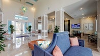This stylish single-floor community space is thoughtfully designed to blend elegance with functionality. The main lounge area immediately impresses with its soaring double-height ceilings, decorative white columns with textured stone panels, and gleaming polished tile flooring. A striking living wall installation anchors one side of the room, paired with a mounted flat-screen display and recessed lighting that creates a warm, inviting atmosphere throughout.

Seating areas are arranged to encourage both relaxation and socializing, featuring rich blue upholstered sofas, tufted leather ottomans, and carved wooden armchairs with coordinating accent pillows. An eye-catching coral-patterned wallcovering adds a coastal, resort-inspired character to one section of the lounge, complemented by wood-look plank flooring and generous natural light from large windows with tailored valances.

Floor-to-ceiling windows in the lounge's brightest corner frame picturesque views of a shimmering outdoor pool surrounded by palm trees, with dramatic drapery framing the outlook. A fully appointed kitchen features white shaker cabinetry, granite countertops, stainless steel appliances including a side-by-side refrigerator, wall oven, and dishwasher, all set against the bold botanical wallcovering. The overall ambiance is fresh, coastal, and modern — a welcoming retreat designed for community enjoyment.