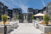 Courtyard 1 in Meridian, Idaho, is a thoughtfully designed outdoor amenity space that blends active recreation with relaxed social gathering. At the heart of the space is a full-size artificial turf sports field, marked with multi-colored lines for soccer and other sports, complete with goals and chain-link fencing. Adjacent to the field, the courtyard opens into a series of well-appointed outdoor living areas that invite both quiet relaxation and lively community interaction. A striking dark-framed pergola with louvered panels serves as the architectural centerpiece, providing partial shade over an expansive arrangement of modern white outdoor furniture, including sofas, benches, and dining tables and chairs. String lights draped overhead lend a warm, inviting ambiance to the space. Adirondack-style chairs and contemporary lounge seating are arranged around fire pits and low-profile planters filled with young trees and greenery. The courtyard is framed on all sides by the multi-story, contemporary residential buildings featuring large windows, private balconies, and a modern façade of mixed materials. Open to the Idaho sky, the space receives abundant natural light, creating a bright, energetic atmosphere suited for year-round outdoor living and community engagement.