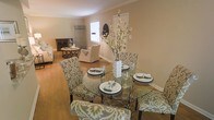 This single-story home showcases an open-concept layout that seamlessly connects the dining and living areas, creating a spacious and inviting atmosphere. The dining area features a round glass-top table surrounded by patterned upholstered chairs in a sage and cream ikat design, adding a stylish focal point to the space. A decorative floral arrangement serves as a centerpiece, complementing the elegant table settings. Crown molding trims the walls throughout, lending a refined architectural detail to the interior. Warm-toned hardwood flooring flows consistently across both areas, enhancing the cohesive feel of the space. The adjacent living area is furnished with neutral-toned seating and accented by decorative pieces, including a wrought-iron side table and a dark wood cabinet that add character and depth. Natural light filters in through a window, brightening the warm beige walls and contributing to the overall airy ambiance. The thoughtful arrangement of furniture and decor throughout creates a balanced, welcoming environment well-suited for both everyday living and entertaining.