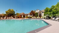 This inviting outdoor amenity space centers around a generously sized swimming pool with crystal-clear turquoise water, designed with a gently curved form that maximizes both swimming area and visual appeal. The pool is framed by a broad, well-maintained concrete sundeck lined with rows of lounge chairs, providing ample space for sunbathing and relaxation. A distinctive covered gazebo with a hip roof anchors one side of the deck, offering a shaded retreat alongside additional outdoor seating with umbrella tables — ideal for casual gathering.

The surrounding landscape is rich with mature trees and manicured plantings, creating a sense of seclusion and natural tranquility. The backdrop features a stately multi-story clubhouse building with a striking white exterior, bold architectural gable detailing, and large glass windows that allow natural light to flow freely. An adjacent single-story structure with a complementary roofline serves as a support facility, likely housing restrooms or changing areas.

The overall ambiance is that of a well-appointed residential community amenity — bright, open, and thoughtfully designed for outdoor leisure. Under clear blue skies, the combination of sparkling water, lush greenery, and classic architectural elements creates an atmosphere that is both functional and aesthetically refined.