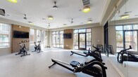 Located at 1835 Morehead Ridge Drive in Charlotte, NC, this well-appointed fitness center offers a spacious, single-level layout designed to support a full range of workout routines. The facility is flooded with natural light through expansive floor-to-ceiling windows and glass-panel doors that line multiple walls, creating an open, energizing atmosphere and offering views of the surrounding outdoor areas, including an accessible deck and pool area.

The fitness center is divided into functional zones. The cardio area features Matrix rowing machines and stationary spin bikes arranged to take full advantage of the bright, airy environment. A dedicated strength and functional training zone houses a well-stocked equipment rack with medicine balls, kettlebells, resistance bands, and stability balls, complemented by additional cycling bikes and a weight bench.

A striking exposed brick accent wall serves as a bold focal point, anchoring a wall-mounted flat-screen TV—ideal for following workout programs or entertainment during training sessions. Full-length mirrors along one wall amplify the sense of space and allow for form monitoring. Ceiling fans and recessed accent lighting with distinctive yellow-green fixtures provide both comfort and style. The durable speckled flooring throughout is practical and easy to maintain, completing this thoughtfully designed, amenity-rich fitness space.