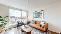 This thoughtfully designed one-bedroom residence combines modern aesthetics with functional living across a single floor. The open-concept layout seamlessly connects the kitchen, dining, and living areas, creating a cohesive and inviting environment. Large windows throughout the space flood every room with natural light while framing sweeping urban views of the surrounding cityscape.

The living area is anchored by a warm tan leather sofa, a sculptural oval coffee table, and a patterned area rug, complemented by curated wall art and a built-in media console. The space opens directly to the kitchen, which features a generous island with a quartz countertop, light wood cabinetry with brushed gold hardware, a black matte pull-down faucet, and stainless steel appliances — ideal for both everyday cooking and entertaining.

The bedroom offers a serene retreat with a large window that welcomes natural light and city views, light wood-tone flooring, and access to a well-appointed walk-in closet. The adjacent bathroom features a light wood vanity with stone countertop, black matte fixtures, and warm wood-look tile flooring. Throughout the residence, wide-plank hardwood-style flooring, clean white walls, and recessed lighting create a cohesive, contemporary ambiance.