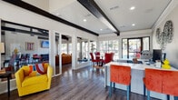 This single-floor communal space makes a striking first impression with its soaring vaulted ceilings adorned with bold, contrasting black exposed beams that run the length of the interior. Rich hardwood flooring flows seamlessly throughout, anchoring the space's warm yet contemporary aesthetic. Large grid-pane windows on multiple walls flood the interior with natural light and offer views of mature trees and outdoor amenities beyond.

The open-concept layout is divided into several functional zones. A reception or workspace area features a sleek white desk with vibrant orange and red accent seating, while an adjacent dining area showcases a round table paired with bold red chairs atop a vivid multicolored area rug. A comfortable lounge area is appointed with a generously sized caramel-toned velvet sofa accented with jewel-toned throw pillows, alongside armchairs and a glass-top coffee table — all centered around a wall-mounted flat-screen TV.

A private room, accessible through glass-paned French doors, offers a dedicated workspace with desks and monitors. The kitchenette features crisp white cabinetry, quartz countertops, a stainless refrigerator, microwave, and coffee station. Vibrant artwork and eclectic décor throughout give this space an energetic, welcoming ambiance suited for gathering, working, or relaxing.