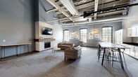 Located at 815 Court St in Utica, New York, this single-floor communal living space masterfully combines industrial heritage with contemporary design. The open-concept layout offers a generous, well-proportioned footprint that feels expansive thanks to dramatically high ceilings with exposed structural beams and large-diameter ductwork—hallmarks of a thoughtfully converted industrial building.

The lounge area serves as the heart of the space, anchored by a striking white tile fireplace surround with a rich wood mantel and a wall-mounted flat-screen television above it. Arched windows along the exposed brick wall flood the seating area with abundant natural light, while curated artwork adds a gallery-like warmth. Comfortable seating, including a leather sofa and accent chairs, is arranged to encourage conversation and relaxation.

Adjacent to the lounge, a well-appointed kitchenette features white shaker cabinetry, dark countertops, a sink, and a dishwasher, complemented by a long high-top bar table with modern stools—ideal for casual dining or working. Near the entry, a welcoming reception area with additional seating and contemporary abstract artwork leads to glass double doors. Linear LED lighting fixtures and patterned carpet tile throughout complete this polished, versatile communal environment.