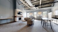 Located at 815 Court St in Utica, New York, this single-floor communal living space masterfully combines industrial heritage with contemporary design. The open-concept layout offers a generous, well-proportioned footprint that feels expansive thanks to dramatically high ceilings with exposed structural beams and large-diameter ductwork—hallmarks of a thoughtfully converted industrial building.

The lounge area serves as the heart of the space, anchored by a striking white tile fireplace surround with a rich wood mantel and a wall-mounted flat-screen television above it. Arched windows along the exposed brick wall flood the seating area with abundant natural light, while curated artwork adds a gallery-like warmth. Comfortable seating, including a leather sofa and accent chairs, is arranged to encourage conversation and relaxation.

Adjacent to the lounge, a well-appointed kitchenette features white shaker cabinetry, dark countertops, a sink, and a dishwasher, complemented by a long high-top bar table with modern stools—ideal for casual dining or working. Near the entry, a welcoming reception area with additional seating and contemporary abstract artwork leads to glass double doors. Linear LED lighting fixtures and patterned carpet tile throughout complete this polished, versatile communal environment.