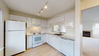 This single-floor residence offers a clean, functional layout across several well-defined living spaces. The open living room is a standout area, featuring vaulted ceilings that lend a sense of spaciousness and airiness, plush neutral carpeting, and a tile-surround fireplace flanked by two windows that draw in generous natural light. The bright, white tones throughout create a calm and welcoming ambiance.

The kitchen is efficiently designed in an L-shaped configuration, outfitted with sleek, high-gloss white cabinetry, gray laminate countertops, and light hardwood-style flooring. A window above the sink provides natural light, while track lighting illuminates the workspace. White appliances — including a refrigerator, electric range, and dishwasher — complete the practical setup.

The bedroom features vaulted ceilings, soft carpeting, and access to an adjacent room, likely a walk-in closet or en-suite. Two bathrooms serve the home: one with a tiled walk-in shower and sliding glass door, and another with a full tub-shower combination and wood-style tile flooring.

A dedicated utility area houses a full-size washer and dryer alongside a water heater, providing convenient in-unit laundry. The overall design emphasizes clean lines, neutral palettes, and practical livability throughout.