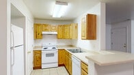This single-floor, one-bedroom, one-bathroom residence offers a practical and cohesive layout across a comfortable footprint. The space includes a hallway, a dedicated closet, and an additional unclassified room, providing versatile utility throughout.

The kitchen is a standout feature, equipped with warm knotty pine cabinetry that extends from base to upper wall cabinets, offering generous storage. White appliances — including an electric range, dishwasher, and refrigerator — complement the light countertops and neutral tile flooring. A built-in skylight panel provides bright overhead illumination, and a peninsula countertop adds a convenient workspace or casual dining area.

The living area is carpeted in a neutral tan tone, creating a warm and inviting atmosphere. Large windows allow natural light to filter in, offering views of surrounding greenery. The space also features a double-door closet, adding practical storage.

The bedroom is carpeted as well, with white walls and a window that brings in natural light and outdoor views. Its modest yet functional size makes it a comfortable private retreat.

The bathroom mirrors the design language of the kitchen, featuring matching knotty pine cabinetry beneath the vanity, a single-sink countertop, a wall-mounted mirror with overhead lighting, and light tile flooring throughout. The overall aesthetic is clean, consistent, and functional.
