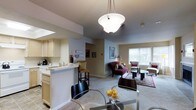 This single-floor residence offers a thoughtfully designed open-concept layout that integrates the kitchen, dining, and living areas into one cohesive and functional living space. The kitchen is outfitted with light wood cabinetry, a double sink, and essential appliances including a coffee maker, all anchored by a breakfast bar with barstool seating — ideal for casual dining or morning routines. A pendant light with a frosted globe shade hangs gracefully above, providing warm, focused illumination. The dining area features a sleek glass-top table surrounded by dark modern chairs, creating an elegant yet understated setting for meals. Flowing seamlessly from the dining space, the living room is generously proportioned and bathed in natural light from large windows dressed with horizontal blinds. The space is furnished with a rich burgundy chaise lounge and accent chairs, arranged around a glass coffee table atop a patterned area rug, lending the room a refined, comfortable character. Framed artwork and soft neutral wall tones throughout contribute to a cohesive and welcoming ambiance. The open sightlines between all areas make the space feel expansive and airy, well-suited for both everyday living and entertaining.