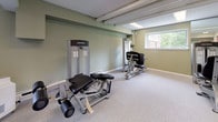This fitness center offers a comprehensive workout facility spread across a single floor, thoughtfully organized into distinct exercise zones. The space is finished with neutral carpet flooring throughout most areas, transitioning to rubber mat flooring in the free weights section for added durability and safety. Sage green walls and recessed fluorescent lighting create a clean, functional atmosphere, while multiple windows allow natural light to filter in and provide views of the surrounding exterior.

The cardio area features stationary bikes, including spin-style cycles, as well as treadmills, all positioned alongside large wall-mounted mirrors that enhance the sense of openness. The strength training zone is well-stocked with Life Fitness selectorized machines, including leg press and leg curl stations, designed for targeted muscle training. A dedicated free weights area houses a full rack of dumbbells alongside benches and weight plates, offering a complete resistance training setup.

Mirrored walls are a prominent design feature throughout, supporting both form-check functionality and visual spaciousness. The facility also includes restroom access, a vending machine, and clearly marked exit doors with distinctive red framing. The layout is open and navigable, providing ample space between equipment for safe, comfortable use.