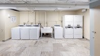 This shared laundry facility offers a practical and well-organized space designed to accommodate the needs of multiple users. The main laundry area is generously sized, housing a row of top-loading washing machines alongside stacked dryer units, providing ample capacity for laundry loads of all sizes. A deep utility sink positioned centrally in the room adds convenience for hand-washing or pre-treating garments. An additional utility sink counter along one wall further expands the workspace available to users. The exposed ceiling features overhead piping and bright fluorescent lighting, giving the space a clean, functional aesthetic. The warm beige walls complement the neutral stone-look tile flooring, which runs throughout the entire facility, lending a polished appearance to an otherwise utilitarian space. Several gray-painted doors lead to additional rooms, including a private restroom. The restroom is compact and efficiently laid out, featuring a toilet, a small bench, a step-on waste bin, and painted concrete block walls that echo the building's sturdy construction. The tile flooring continues seamlessly into this area. Overall, this laundry room is thoughtfully outfitted to deliver a complete, convenient, and clean laundering experience.