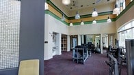 This fitness center makes a strong impression with its dramatically high ceilings, creating an open and energizing atmosphere for workouts. Decorative horizontal striping in green, yellow, and red encircles the upper walls, adding a bold visual element that distinguishes the space. A series of industrial-style pendant lights hang at varying heights, providing ample illumination throughout the area.

The workout floor is covered in durable, dark patterned carpeting and is equipped with a variety of strength-training machines, a well-stocked dumbbell rack, and cardio equipment. Large mirrors line one wall, enhancing the sense of space and allowing users to monitor their form. Natural light filters in through windows visible toward the rear of the space, contributing to a bright and welcoming environment.

Additional functional features include built-in open shelving and a water fountain station tucked into a recessed alcove along one wall, offering convenient access to storage and hydration. The overall layout is thoughtfully organized, balancing equipment density with open floor space to ensure a comfortable and practical fitness experience.