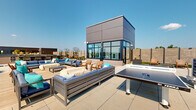 Located at 713 Lamont Street Northwest in Washington, DC, this rooftop terrace offers a thoughtfully designed outdoor amenity space with a wide range of recreational and social features. The terrace spans a generous footprint and is organized into several distinct zones, each serving a different function while contributing to a cohesive, modern aesthetic.

At the heart of the space, a central glazed pavilion structure with dark paneled cladding and floor-to-ceiling windows serves as an architectural focal point. Surrounding it, multiple seating arrangements feature plush, blue-cushioned sectional sofas, lounge chairs, and wood-accented furniture, creating inviting areas for relaxation and socializing. A dedicated deck area with warm wood composite flooring provides an intimate lounge setting adjacent to the pavilion.

For recreation, a full-size ping-pong table and a cornhole board are positioned on the open patio areas, while bar-height seating and a grill station offer options for outdoor dining and entertaining. Sun loungers are arranged in quieter corners, ideal for unwinding.

Lush landscaping, including ornamental grasses, low shrubs, and planted beds framed by horizontal slatted privacy screens, adds greenery and visual texture throughout. The open-sky setting allows panoramic views of the surrounding Washington, DC cityscape, making this rooftop terrace a vibrant and functional outdoor retreat.