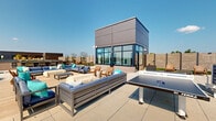 Located at 713 Lamont Street Northwest in Washington, DC, this rooftop terrace offers a thoughtfully designed outdoor amenity space with a wide range of recreational and social features. The terrace spans a generous footprint and is organized into several distinct zones, each serving a different function while contributing to a cohesive, modern aesthetic.

At the heart of the space, a central glazed pavilion structure with dark paneled cladding and floor-to-ceiling windows serves as an architectural focal point. Surrounding it, multiple seating arrangements feature plush, blue-cushioned sectional sofas, lounge chairs, and wood-accented furniture, creating inviting areas for relaxation and socializing. A dedicated deck area with warm wood composite flooring provides an intimate lounge setting adjacent to the pavilion.

For recreation, a full-size ping-pong table and a cornhole board are positioned on the open patio areas, while bar-height seating and a grill station offer options for outdoor dining and entertaining. Sun loungers are arranged in quieter corners, ideal for unwinding.

Lush landscaping, including ornamental grasses, low shrubs, and planted beds framed by horizontal slatted privacy screens, adds greenery and visual texture throughout. The open-sky setting allows panoramic views of the surrounding Washington, DC cityscape, making this rooftop terrace a vibrant and functional outdoor retreat.