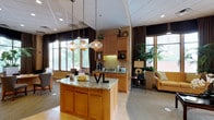 This single-floor leasing office is thoughtfully designed to create a warm and professional first impression for prospective residents. The entry foyer greets visitors with light hardwood flooring, a curved reception desk finished in light wood with decorative lattice paneling, and tasteful artwork adorning the walls. Signage and decorative accents lend a branded, polished feel to the arrival experience.

The main open area transitions seamlessly from the reception zone into a spacious lounge and hospitality area. A central island with rich wood cabinetry and a granite countertop serves as a natural focal point, illuminated by a cluster of elegant glass pendant lights overhead. Adjacent to the island, warm honey-toned cabinetry houses a beverage station equipped with a coffee maker and sink, adding a functional and welcoming touch for visitors.

Large windows dressed with rich, dark draping allow abundant natural light to fill the space while maintaining a refined aesthetic with views of lush greenery outdoors. The lounge area features comfortable seating arrangements, including upholstered chairs and a sofa, arranged around a carpeted area rug. A dedicated meeting table with cushioned chairs provides a functional space for consultations. Tall indoor plants add organic warmth throughout, contributing to the overall inviting and professional ambiance of the office.
