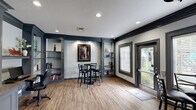 This single-floor business center in Baton Rouge, Louisiana, is designed with a refined, professional aesthetic that balances functionality with sophisticated style. The interior features a consistent color palette of light gray walls contrasted by bold dark charcoal trim, crown molding, and framed doorways, lending the space a sharp, polished character throughout.

The workspace area is anchored by an expansive built-in wall unit finished in charcoal gray, incorporating floor-to-ceiling shelving, cabinetry, and a built-in desk surface accommodating dual workstations. Recessed lighting illuminates the shelving, which provides ample display and storage space. Wood-look plank flooring runs throughout, adding warmth to the otherwise cool-toned design.

Multiple bistro-style seating areas with round tables and ladder-back chairs offer comfortable spots for informal meetings or casual work sessions. Open shelving units with decorative accents are strategically placed throughout, contributing both visual interest and practical storage.

Natural light filters into the space through several windows fitted with horizontal blinds, while a glass-panel entry door opens directly to an outdoor area, further brightening the interior. Recessed ceiling fixtures provide consistent overhead illumination, ensuring a well-lit environment at all times. Overall, the space delivers a functional yet stylish setting well-suited for professional productivity.