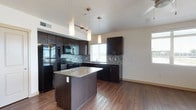 This single-floor residence offers a thoughtfully designed open-concept layout that seamlessly connects the kitchen and living areas, creating a spacious and functional environment. The kitchen is a standout feature, showcasing rich dark wood cabinetry that extends to the upper walls, providing generous storage. A central island topped with light granite countertops serves as both a prep space and a casual gathering point. Matching granite lines the perimeter countertops, complemented by a stylish mosaic tile backsplash that adds texture and visual interest. Black appliances, including a refrigerator, range, and built-in microwave, lend a sleek, cohesive aesthetic. Pendant lighting fixtures hang above the island, adding warmth and focused illumination, while a track lighting system further brightens the kitchen area. Warm hardwood-style flooring runs throughout, grounding the space with a welcoming, contemporary feel. Windows along multiple walls invite natural light deep into the interior, enhancing the open and airy ambiance. The textured ceiling adds a subtle architectural detail that completes the polished look of this well-appointed residence.