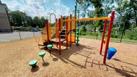 This community space is thoughtfully designed to offer a blend of recreational and functional amenities across a single level. The outdoor playground area is a standout feature, showcasing a colorful multi-level play structure finished in bold red, yellow, and orange. The play equipment includes climbing elements, stairs, a slide, and spiral pole features, all set atop a generous wood chip safety surface. Freestanding green spinning seats and activity panels are positioned throughout the area, encouraging active play. The playground is enclosed by a chain-link fence and framed by mature trees, providing a natural backdrop and partial shade.

Inside, the business center is a dedicated, well-appointed workspace designed for productivity. It features an L-shaped granite-topped workstation with multiple desktop computers and ergonomic task chairs, making it suitable for individual or concurrent use. A separate area houses a multifunction printer and additional counter space for printing and copying needs. The rooms are finished with warm beige walls, crown molding, and carpet flooring, lending a polished, professional feel. Glass-paneled French doors provide both privacy and visual openness, connecting the business center to the broader interior common areas, which include a welcoming foyer with decorative furnishings and light hardwood flooring.