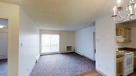 Located at 600 South Dayton Street in Denver, Colorado, this single-floor unit offers a straightforward and functional layout designed for comfortable everyday living. The living area serves as the heart of the space, featuring plush carpeting that transitions to hard flooring near the entryway, providing both warmth and practicality underfoot. A large sliding window dominates one wall, flooding the room with natural light and offering a view to an outdoor balcony area. Vertical blinds provide adjustable privacy and light control. A wall-mounted air conditioning unit complements the baseboard heating system that runs along the perimeter of the room, ensuring year-round climate comfort. The textured popcorn ceiling adds a classic residential character to the space. The clean, neutral-toned walls create a versatile backdrop suitable for a variety of interior styles. A doorway visible from the main living area leads further into the unit, connecting to additional rooms. The overall ambiance is bright, open, and move-in ready, making it a practical choice for those seeking a well-lit, low-maintenance living space in the Denver area.