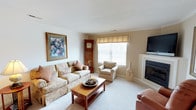 This single-floor space centers around a well-appointed living room designed for comfort and functionality. The room features neutral beige walls and plush carpeting that contribute to its warm, inviting atmosphere. A fireplace with a clean white mantel serves as a natural focal point on one wall, complemented by a wall-mounted flat-screen television above. Natural light flows in through a window dressed with a Roman shade and horizontal blinds, brightening the space throughout the day. The seating arrangement includes a patterned sofa and coordinating armchair in soft, muted tones, anchored by a wooden coffee table with a lower shelf for additional storage. A tall grandfather clock stands in the corner, adding a classic, timeless element to the room's décor. Decorative touches, including a framed floral artwork and ceramic vases, enhance the traditional aesthetic. The overall ambiance is calm and understated, with a cohesive color scheme of creams, tans, and soft earth tones throughout.