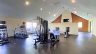 This single-floor fitness center is designed to support a comprehensive workout experience. The space is anchored by vaulted ceilings that lend an open, airy feel, complemented by recessed lighting and a ceiling fan that ensure comfortable conditions during exercise. The room features durable, dark rubber flooring ideal for a high-activity environment.

At the center of the space stands a multi-station strength training machine, providing a wide range of resistance exercises for users of all fitness levels. Surrounding it are several cardio machines, including what appear to be an elliptical trainer, a stair climber, and an upright exercise bike, offering diverse options for cardiovascular workouts.

Natural light filters in through a window on one wall, brightening the space and offering a pleasant view of greenery outside. A wall-mounted flat-screen television is positioned at the far end of the room, allowing users to stay entertained during their sessions. An accent wall painted in a warm terracotta tone adds a subtle visual interest to the otherwise neutral color palette. The overall layout is thoughtfully organized, providing ample space to move between equipment safely and comfortably.