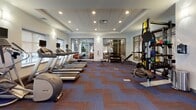 Located at 100 Exchange St in Malden, Massachusetts, this modern fitness center offers a comprehensive workout environment designed to support a wide range of exercise routines. The space is laid out on a single level and features a distinctive blue and brown chevron-patterned carpet throughout, lending a polished, contemporary aesthetic to the interior.

The cardio zone is well-appointed with multiple treadmills, elliptical trainers, and upright bikes, each equipped with individual mounted flat-screen monitors for an engaging workout experience. Large windows along the perimeter allow natural light to fill the space while providing views of the surrounding area. Recessed lighting and a coffered ceiling add architectural refinement overhead.

A dedicated strength and functional training area includes a multi-use cable machine, a power rack stocked with TRX straps, battle ropes, and resistance bands, as well as a full rack of medicine balls in varying weights. Adjustable benches and free weights complete the strength training offerings. Glass-paneled interior walls connect different zones of the facility, maintaining an open, airy feel while defining distinct workout areas. The space also includes a separate room visible through glass partitions, and a private door for additional utility. Sanitizing stations are conveniently positioned near the entrance, reflecting a clean and well-maintained facility.