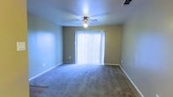 This single-floor space features a open living area finished with wall-to-wall carpeting in a neutral gray tone, providing a comfortable and cohesive foundation throughout. The room is dressed in soft, muted green-gray walls that contribute to a calm and understated ambiance. A ceiling fan with an integrated light fixture is centrally mounted, offering both air circulation and ambient lighting. A large sliding glass door dominates one wall, dressed with full-length vertical blinds that diffuse natural light evenly across the room, brightening the interior while maintaining privacy. The door suggests potential access to an outdoor area. Practical details include visible HVAC ceiling vents for climate control, standard white baseboards framing the perimeter of the room, and conveniently placed electrical outlets along the walls. The overall layout is open and unfurnished, allowing for flexible arrangement to suit a variety of uses. The space presents a clean, move-in ready environment with functional infrastructure already in place.