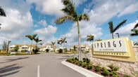 Springs at Traditions is a residential apartment community characterized by a polished, Florida-inspired aesthetic. The property makes a strong first impression with its grand entrance drive, flanked by tall swaying palm trees and colorful, manicured tropical landscaping including ornamental grasses and flowering shrubs set against rich mulch beds. A prominent stone monument sign marks the community entrance, reflecting a sense of permanence and quality.

The buildings visible from the entry are designed in a coastal vernacular style, featuring neutral earth-toned exteriors, pitched rooflines, and architectural detailing consistent with upscale Florida residential construction. The community is laid out across a single story, suggesting a spread-out, accessible campus that prioritizes ease of movement throughout the grounds.

The wide, well-paved entry road provides clear circulation through the community, contributing to an open and organized feel. An American flag is visible near the leasing or community center building, which appears prominently positioned at the heart of the property.

Overall, the community presents a bright, resort-like atmosphere enhanced by abundant natural light, open skies, and lush greenery that is characteristic of Southwest Florida living. The landscaping and architectural cohesion combine to create an inviting, well-curated residential environment.