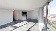 This single-floor space presents a sleek, modern design characterized by an open-concept layout that seamlessly connects the living area to the kitchen. The expansive main living area is finished with wide-plank hardwood-style flooring in a cool gray tone, lending a clean and contemporary aesthetic throughout. Large windows along one wall flood the interior with abundant natural light, casting dramatic sunbeams across the floor and creating a bright, airy atmosphere.

The kitchen is positioned at the rear of the open floor plan and features dark cabinetry that provides a striking contrast against the light gray walls. A peninsula counter serves as a natural divider between the kitchen and the living space, offering both additional workspace and a visual anchor to the layout. Track lighting installed on the kitchen ceiling ensures the workspace is well-illuminated. Stainless steel appliances, including a microwave and range, add a polished, modern finish to the culinary space.

The neutral gray walls and white ceilings throughout contribute to a cohesive, minimalist palette that allows for versatile interior styling. A climate control unit is integrated along the base of the window wall, ensuring year-round comfort. The overall ambiance is clean, open, and move-in ready, with thoughtful design details that balance style and practicality.