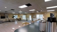 Located on Hearthside Drive in Wilmington, North Carolina, this single-story space offers a spacious, open interior designed for comfort and community use. The layout features a drop ceiling with recessed panel lighting and skylight-style flush fixtures that provide consistent, even illumination throughout. Ceiling fans are evenly distributed across the space, ensuring airflow and comfort year-round.

Large windows line multiple walls, inviting generous natural light and offering views of the surrounding greenery outside. The warm beige walls and white wainscoting trim create a neutral, inviting palette throughout. The flooring transitions between polished tile in the central area and carpet in the seating zones, defining distinct functional areas within the open floor plan.

A front counter or reception surface with a dark finish anchors one side of the space, suited for administrative or service use. Multiple seating arrangements with upholstered chairs are positioned near the windows, creating relaxed conversation areas. Decorative framed artwork and accent lamps add warmth and a residential feel to the overall environment, making the space feel approachable and well-maintained.