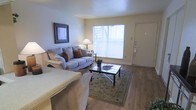 This single-story space centers around a welcoming living area finished with warm hardwood-style flooring that extends throughout the room. A large window along the main wall allows natural light to fill the space, creating a bright and comfortable atmosphere. The living area is well-proportioned, offering enough room for a plush sofa, accent chairs, and a glass-topped coffee table resting on a decorative area rug. Neutral wall tones complement the light flooring, giving the space a clean and cohesive look. A framed piece of wall art adds a decorative touch, while a floor lamp provides supplemental ambient lighting. The front entry door is positioned adjacent to the living area, creating a straightforward and functional layout that connects the main entrance directly to the primary living space. A partial view of an additional seating area or adjoining room is visible, suggesting a practical open flow between spaces. Overall, the living area conveys a modest, well-maintained environment with a neutral palette and functional design suited for everyday living.
