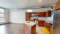 This single-floor unit offers a thoughtfully designed open-concept layout that seamlessly connects the kitchen and living areas. The kitchen is a standout feature, showcasing rich warm-toned wood cabinetry, granite countertops, and a full suite of stainless steel appliances, including a gas range, built-in microwave, and refrigerator. A curved kitchen island with a pendant light above serves as a natural focal point, providing additional workspace and casual dining potential. Classic subway tile backsplash adds a clean, timeless touch, while tile flooring grounds the cooking space. The living area transitions smoothly via durable wood-look flooring and is bathed in natural light through expansive floor-to-ceiling windows fitted with horizontal blinds. These large windows frame views of the surrounding urban landscape, lending an airy, open ambiance throughout. An exposed metallic duct runs along the ceiling, contributing a distinct industrial-modern aesthetic. The bathroom is well-appointed with a wood-toned vanity, granite countertop, an undermount sink, and a full shower-tub combination. Throughout the unit, crisp white walls provide a neutral backdrop that complements the warm wood finishes, creating a balanced and welcoming interior.