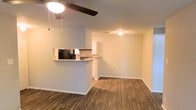 This single-floor space offers a practical and comfortable layout centered around an open-concept living and kitchen area. The living space is outfitted with rich, warm hardwood-style flooring that extends throughout, providing a cohesive and inviting atmosphere. A ceiling fan with a light fixture is positioned centrally overhead, offering both air circulation and ambient lighting.

The kitchen is defined by a raised breakfast bar counter that serves as a natural divider between the cooking and living areas while maintaining the open feel of the space. White cabinetry lines the kitchen walls, providing ample storage, and built-in appliances — including a microwave — are integrated into the design. The layout offers functional workspace while keeping the area visually connected to the rest of the floor plan.

Neutral, light gray walls complement the warm tones of the flooring and cabinetry, creating a balanced and understated aesthetic throughout. Recessed ceiling fixtures provide additional lighting in both the living and kitchen zones. White baseboards trim the perimeter cleanly, adding a crisp, finished detail to the overall interior. The single-floor design makes the space straightforward to navigate, with a layout well-suited for everyday living and entertaining.