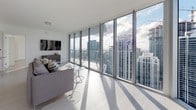 Unit 49M is a thoughtfully designed high-rise residence that combines contemporary architecture with refined finishes throughout. The open-concept living area is an immediate standout, featuring expansive floor-to-ceiling glass windows and sliding doors that flood the space with natural light and frame spectacular panoramic views of the surrounding city skyline. Light-toned hardwood flooring runs throughout the main living areas, enhancing the bright and airy ambiance.

The kitchen is a centerpiece of modern design, outfitted with sleek high-gloss white cabinetry, dark quartz countertops, a generous central island, and a full suite of stainless steel appliances including a Bosch oven, built-in microwave, French door refrigerator, and a wine cooler. Pendant lighting and recessed fixtures provide both functional and ambient illumination.

The bedrooms offer generous proportions, with large sliding glass doors leading to outdoor access and commanding views of the cityscape. The bathrooms are finished to a high standard, featuring striking marble-patterned tile flooring, white cabinetry with chrome hardware, and glass-enclosed shower and tub combinations. A powder room with matching marble tile and a louvered storage cabinet provides additional convenience. The overall aesthetic is clean, modern, and sophisticated throughout.