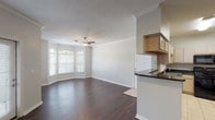 This single-story, one-bedroom, one-bathroom unit offers a thoughtfully organized layout with distinct living, kitchen, and sleeping areas. The living room serves as the centerpiece of the space, showcasing rich, dark hardwood flooring, crown molding, and a ceiling fan with light fixture. A distinctive bay window arrangement allows natural light to filter in, creating a bright and welcoming atmosphere. The open layout flows seamlessly from the living area toward the kitchen and bedroom corridor.

The kitchen is well-equipped with light wood cabinetry, dark granite-style countertops, and a full suite of black appliances, including a refrigerator, dishwasher, and range. Light tile flooring contrasts with the darker counters, and a pass-through counter connects the kitchen to the living area. The bathroom continues the consistent design palette with matching light wood cabinetry, a dark granite vanity top, a large wall mirror with overhead lighting, and tile flooring.

The bedroom offers plush carpeting, a ceiling fan, and a window that brings in natural light, creating a comfortable and restful environment. Overall, the unit presents a cohesive, neutral aesthetic with practical finishes throughout.